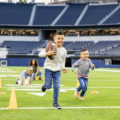 Children playing football on a stadium field, running near orange cones with a football in hand, and one crouching behind.
