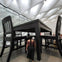 People walking under an oversized table and chairs in a modern exhibition space with a high, white ceiling.