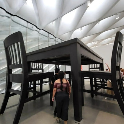 People walking under an oversized table and chairs in a modern exhibition space with a high, white ceiling.