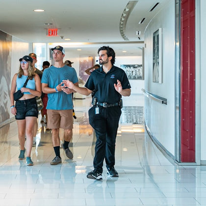 A guide gestures while leading a group of visitors through a bright hallway with glossy flooring and wall decor.