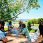 A group of people sitting outdoors at a wooden table, enjoying drinks with a scenic landscape of trees and a pond in the background.