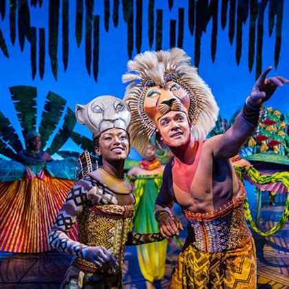 Performers in colorful costumes and lion masks on stage with a jungle-themed backdrop.