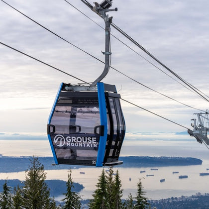 A Grouse Mountain cable car suspended above a scenic landscape with trees, water, and distant ships and islands.