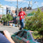 A man in a red shirt and jeans stands on a creatively painted car surrounded by greenery on a city street.