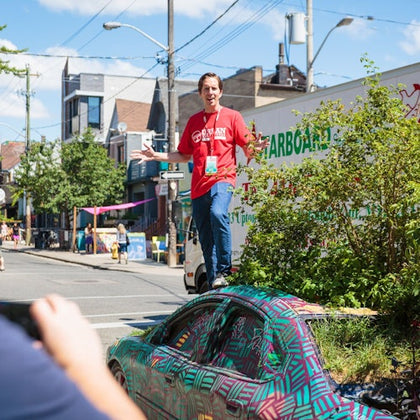 A man in a red shirt and jeans stands on a creatively painted car surrounded by greenery on a city street.