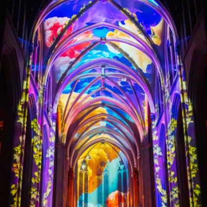 A cathedral interior lit with vibrant, colorful projections of clouds and abstract patterns on the arches and ceiling.