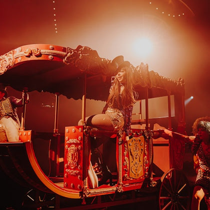 Performer seated on a vibrant, ornate carriage under bright stage lighting, with supporting cast in theatrical costumes.