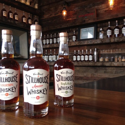 Three bottles of Stillhouse whiskey on a wooden counter in a rustic bar setting with shelves of bottles in the background.