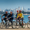 A group of cyclists wearing helmets ride along a waterfront path with a city skyline in the background.