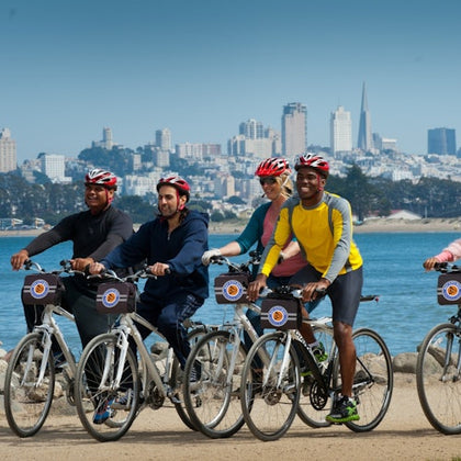 A group of cyclists wearing helmets ride along a waterfront path with a city skyline in the background.