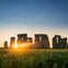 Stonehenge at sunrise with the sun casting light through the stones, set on a green field under a clear sky.