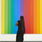 A person with long hair stands in front of a vertical rainbow-striped artwork, observing it thoughtfully in a gallery.