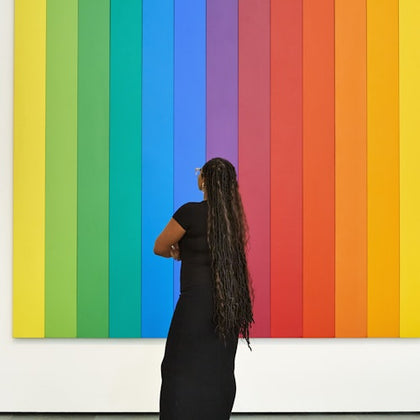 A person with long hair stands in front of a vertical rainbow-striped artwork, observing it thoughtfully in a gallery.