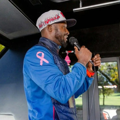 A man in a blue jacket and baseball cap speaks into a microphone inside a vehicle with a window showing greenery outside.