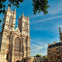 A historic Gothic-style cathedral with twin towers under a blue sky, surrounded by trees and adjacent buildings.