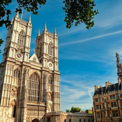 A historic Gothic-style cathedral with twin towers under a blue sky, surrounded by trees and adjacent buildings.