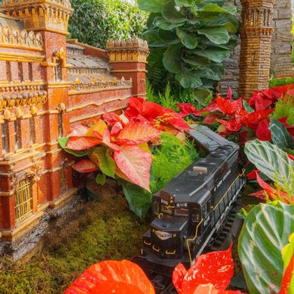 A detailed model of a train passing by a miniature building surrounded by vibrant red and green foliage.