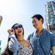 A smiling couple, the woman in sunglasses, stands in front of a towering sign that reads "UNIVERSAL," on a clear, sunny day.
