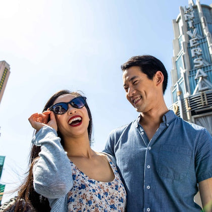 A smiling couple, the woman in sunglasses, stands in front of a towering sign that reads "UNIVERSAL," on a clear, sunny day.