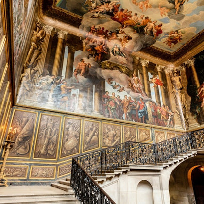 Grand staircase with ornate wrought iron railing, beneath elaborately painted walls and ceiling depicting historical and mythological scenes.