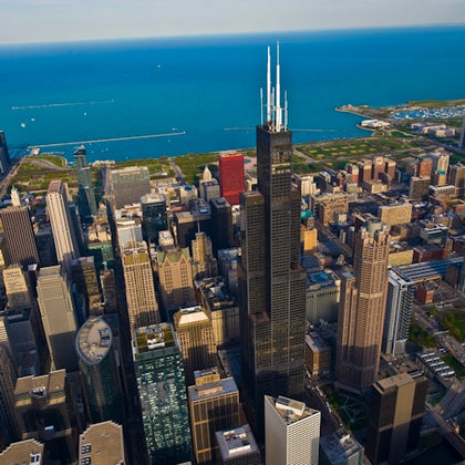 Aerial view of a cityscape with modern skyscrapers, green spaces, and a large body of water in the background.