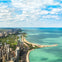 Aerial view of a city with tall buildings along a coastline, featuring beaches, green parks, and a vast blue lake beneath a partly cloudy sky.