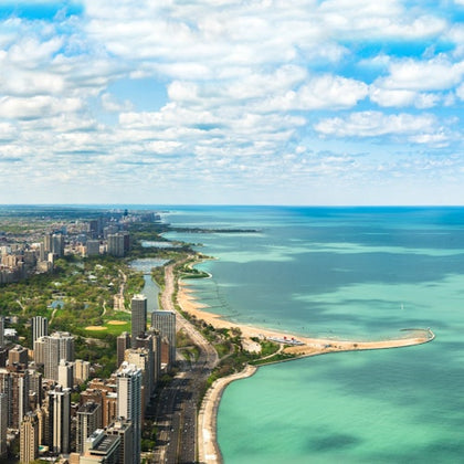Aerial view of a city with tall buildings along a coastline, featuring beaches, green parks, and a vast blue lake beneath a partly cloudy sky.
