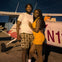 A smiling couple stands in front of a small white and red airplane at night. The man wears a white shirt and checkered shorts, and the woman wears an orange dress.