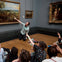 A man kneels and points at a large painting in an art gallery while a group of seated people listens and observes.