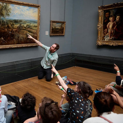 A man kneels and points at a large painting in an art gallery while a group of seated people listens and observes.