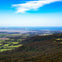A panoramic view of a lush valley with forests and fields, a distant town, and the sea under a blue sky with clouds.