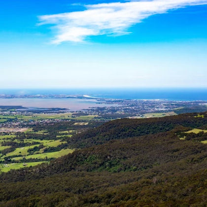 A panoramic view of a lush valley with forests and fields, a distant town, and the sea under a blue sky with clouds.