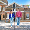 A couple holds hands while walking on a cobblestone path in front of a historic brick building under a clear blue sky.