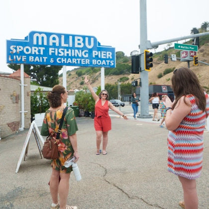 Three women near the Malibu Sport Fishing Pier sign, one posing for a photo while another takes the picture in a casual setting.