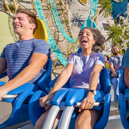 Two people smiling and screaming with excitement while riding a roller coaster against a sunny backdrop.