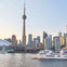 A cityscape view of Toronto with the CN Tower, skyscrapers, and boats on the waterfront under a clear sky.