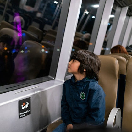 A child in a blue jacket looks out a window while sitting on a train at night.