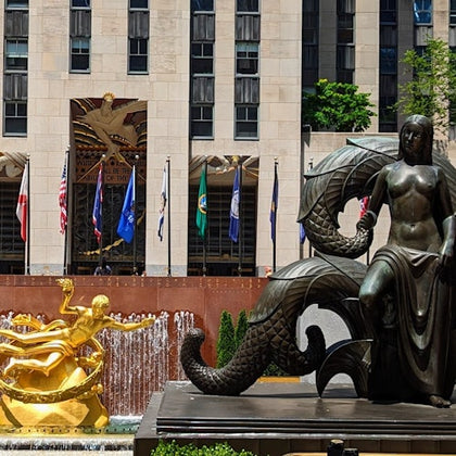 A mermaid statue and a golden Prometheus statue with fountains in a plaza surrounded by flags and a building facade.