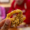 A hand holding a piece of fried calamari, with a blurred background showing a person in a red outfit.