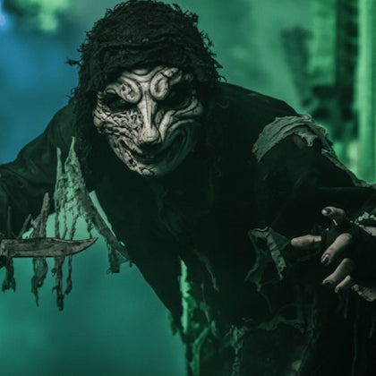 A person in eerie, tattered clothing and a grotesque mask holding a knife, surrounded by greenish fog.