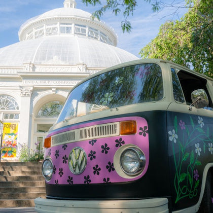 A vintage Volkswagen van with floral designs parked in front of the Enid A. Haupt Conservatory on a sunny day.
