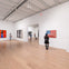People observe modern art pieces in a spacious, white-walled gallery with wooden floors and a high ceiling.