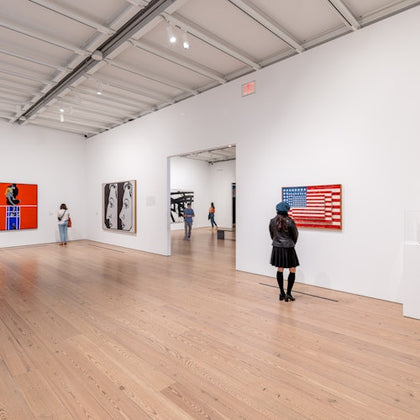People observe modern art pieces in a spacious, white-walled gallery with wooden floors and a high ceiling.