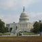 The United States Capitol building with its dome, surrounded by trees, statues, and a lawn under a partly cloudy sky.