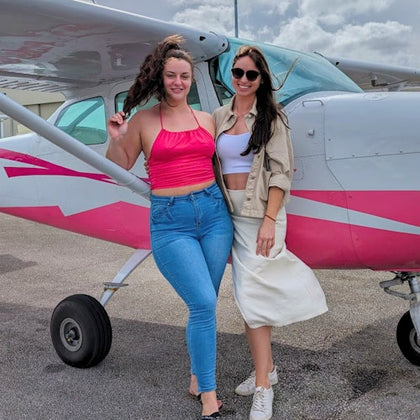 Two women stand smiling in front of a small pink and white airplane on a tarmac.