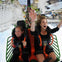 Two girls scream with excitement, holding onto safety restraints on a rollercoaster, surrounded by other riders and amusement park structures.