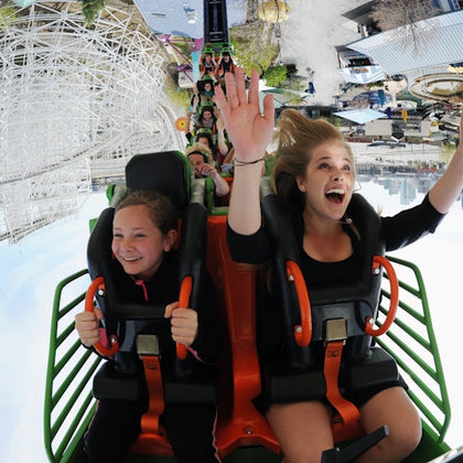 Two girls scream with excitement, holding onto safety restraints on a rollercoaster, surrounded by other riders and amusement park structures.