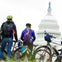 Three cyclists with helmets and backpacks stand with their bikes, facing the United States Capitol dome.