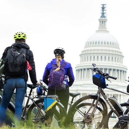 Three cyclists with helmets and backpacks stand with their bikes, facing the United States Capitol dome.
