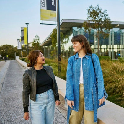 Two women walking and smiling on a pathway next to a modern building and greenery. One wears a denim jacket; the other, a black jacket.
